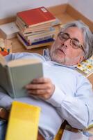 Middle-aged man engages in reading an interesting book lying on the floor full of books. photo