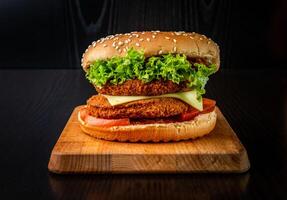 Double Chicken Burger with Cheese, Lettuce, and Tomato isolated on wooden board side view of fastfood on dark background photo