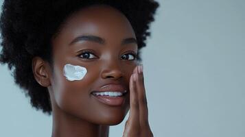 Advertisement image of a Black woman with flawless skin, softly smiling as she applies a moisturizing cream to her cheek with her fingertips, set against a clean, neutral gray background, perfect photo