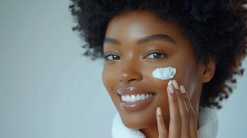 Advertisement image of a Black woman with flawless skin, softly smiling as she applies a moisturizing cream to her cheek with her fingertips, set against a clean, neutral gray background, perfect photo