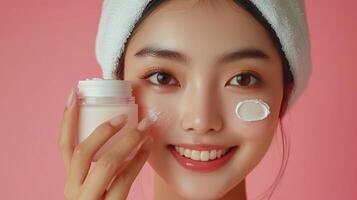 Commercial-style close-up of a young Southeast Asian woman with a bright smile, her hair wrapped in a white towel, holding a jar of cream over one eye while dabs of cream are applied to her photo