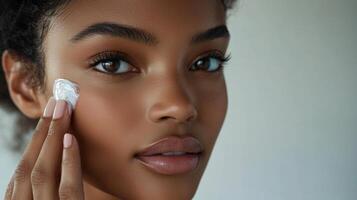 In a bright, minimalist setting, a close-up of a Black woman with dark brown hair and clear skin, carefully applying cream to her face, her calm expression enhancing the appeal of the skincare photo