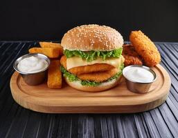 Homemade fish burger with white cream source, cheddar cheese and vegetables with fired fish on serving wooden board on black background. photo