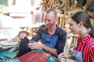 Young female leather maker with craftsman looking and checking the neatness of the leathers bag before sell to customer in workshop. photo