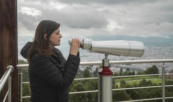 woman, standing at a viewing platform, holding in her hands long binoculars mounted on a metal pedestal against a cloudy sky background photo