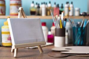 small wooden easel with blank canvas on wooden table against background of shelves with paint tubes, brushes and pencils photo