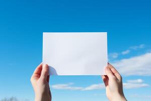 Hands holding a blank white piece of paper in front of a blue sky with white clouds. The image is simple and clean, with plenty of space for text. photo
