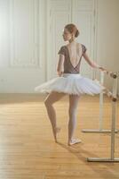 Young ballerina in tutu skirt stretching practicing ballet positions dancing near ballet barre in dance class. Young classical dancer woman in dance studio. Ballet class training Ballet school concept photo