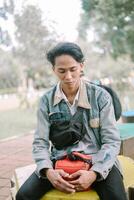 A Southeast Asian man wearing a denim jacket, sitting on a bench in a city square, photo