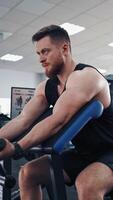Side view of an athletic man during workout in a gym. Bodybuilder sitting in a modern simulator and trains his muscles of arms. Vertical video