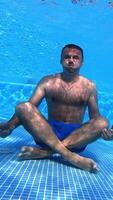Man sits on the bottom of swimming pool. Healthy man is sitting in yoga pose underwater on blue background. Underwater sport. Vertical video