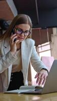 Beautiful working lady standing at the desk with laptop. Busy woman speaking on the phone and checking her notes. Office background. Vertical video