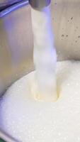 Fresh milk is pouring into a metal bucket creating foam on top. Close up. Food factory production concept. Vertical video