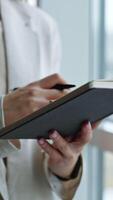 Unrecognizable woman holding a notebook in one hand and pen in another. Lady is taking notes into her book. Close up. Vertical video