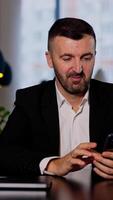 Calm smiling man sitting at table looking at his phone. Businessman is texting message to someone in a big office. Blurred backdrop. Vertical video