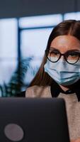 Focused lady in mask works at her laptop in office. Businesswoman closes laptop, puts off her glasses and mask putting them on computer. Vertical video