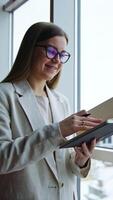 Smiling young woman writing something down in her book. Taking notes while standing a t the window. Cityscape in blur at the backdrop. Vertical video
