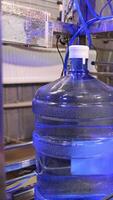 Big full bottles of clean water move by the conveyor belt. Machine is lifting a bottle and worker covers it in plastic bag. Vertical video