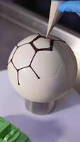 Making cake in a shape of soccer ball. Confectioner's hands applying chocolate lines on the spherical cake. Close up. Vertical video
