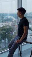 Young smiling man stands in office leaning on desk. Thoughtful male employee looks into panoramic window and then into camera. Cityscape at backdrop. Vertical video