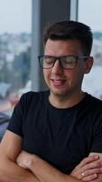 Smiling dark-haired young man puts off his glasses. Businessman standing in empty office thoughtfully looking through the window. Vertical video