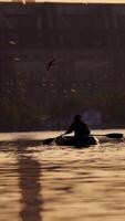 Old man in inflatable boat sailing in the evening. Fisherman in a rowboat with two oars on the background of a big plant. Man tries to find a good place for fishing in the river. Vertical video