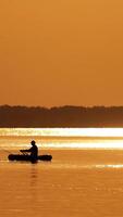 Fisherman in a boat at golden sunset time. Silhouette of man floating with oars on a small rowboat in the evening. Beautiful sunset in the river and man in boat with fishing rods. Vertical video