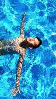 Young boy in the swimming pool. Teenager lying on his back on the water surface in a sunny day. Blue clear water of a pool and a boy swimming. Vertical video