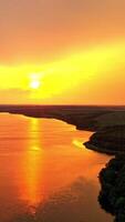 Beautiful sunset near the river in summer. Aerial view of evening environment of calm river among green fields and factory. Camera moves up. Vertical video