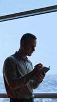 Thoughtful male office worker writes something in his notebook. Adult man taking notes standing at the window. Cityscape at backdrop. Vertical video