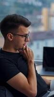 Pondering work issues sitting in the office. Young male office employee sitting in bean bag chair looking into window. Man writing down ideas into paper notebook. Blurred backdrop. Vertical video