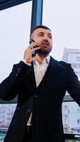 Confident experienced businessman having business conversation. Talking man stands near the window facing office. Low angle view. Vertical video