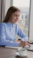 Young businesswoman sitting at desk writing down something into her paper notebook. Panoramic windows with cityscape in blur at backdrop. Vertical video