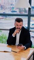 Thoughtful businessman sitting at the table holding phone in hand. The notebook and pen are in front of him. Cityscape at backdrop in windows. Vertical video