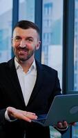 Smiling cheerful bearded man in official suit holds laptop in hands. Man speaks to the camera standing at the panoramic windows. Vertical video