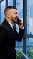Businessman in suit speaks on the phone cheerfully standing near the big window in office. Man talks. Smiles and looks into the window. Vertical video