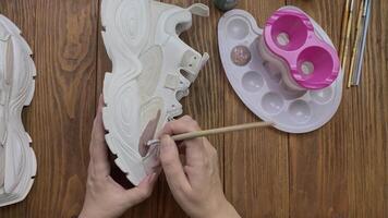 Hands drawing on white boots with brush on desktop. Palette, acrylic paint, bottle with water and other tools on wooden table. Designer's workshop. Hobby, creativity and handmade work at home. video