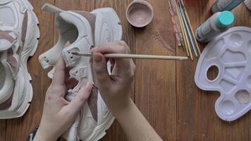 Flat lay of women's hands paint white boots with brush on wooden table. Palette with brown acrylic and other tools on desktop. Designer's workshop. Hobbies, creativity and handmade work at home. video