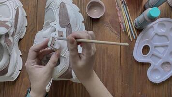 Flat lay of girl's hands paint sneakers with brush on wooden table. Palette with brown acrylic and other tools on desktop. Hobbies, creativity and handmade work at home. Designer's workshop. video