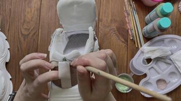 Person's hands paint sneakers with brush on wooden table. Designer's workshop. Palette with brown and green acrylic and other tools on desktop. Hobbies, creativity and handmade work at home. video