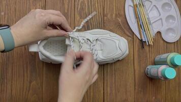 Top view of women's hands pulling out shoelace from sports sneaker. White boot, palette, bottle with paint and brushes on wooden table. video