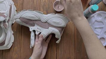 Female's hands dip brush in brown paint and apply it to boot. Palette, tools and tubes on wooden table. Design of sports shoes using acrylic. Hobbies, manual work and leisure at home. video