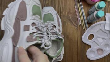 Person's hands take colored sneakers and twist it in front of camera. Plastic palette, acrylic paints, brushes and napkins on wooden table. Shoe design, hobbies and leisure at home. video