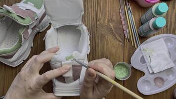 Top view of girl's hands paint sneakers with brush on wooden table. Designer's workshop. Palette with green acrylic and other tools on desktop. Hobbies, creativity and handmade work at home. video
