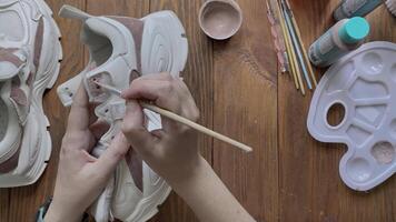 Top view of women's hands paint white boots with brush on wooden table. Palette with brown acrylic and other tools on desktop. Hobbies, creativity and handmade work at home. Designer's workshop. video