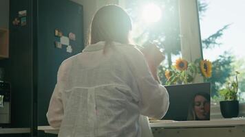 Woman is working remotely at desk in home office, taking notes in notebook. Back view of female freelancer using laptop in the morning video