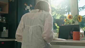 Woman is working remotely at desk in home office, taking notes in notebook. Back view of female freelancer using laptop in the morning video