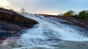 waterfall flowing over smooth, rocky terrain surrounded by lush green forest, The crystal-clear water cascades gently into a calm river below, creating a peaceful and natural, landscape video
