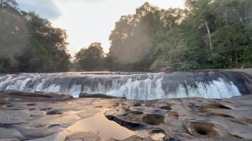 waterfall flowing over smooth, rocky terrain surrounded by lush green forest, The crystal-clear water cascades gently into a calm river below, creating a peaceful and natural, landscape video