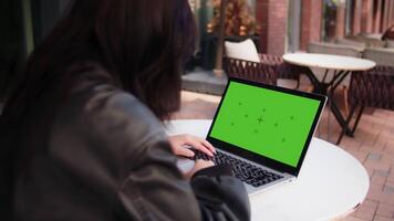 Young girl watching on a laptop with a mockup green screen. Woman in a black jacket is sitting at the outdoor cafe and scrolling feed on a computer, back view video